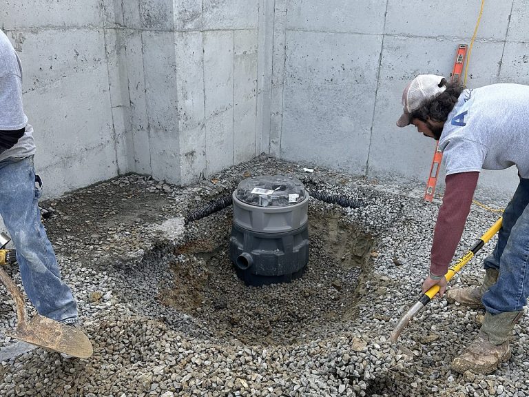 Two plumbers setting a basin in a gravel-filled concrete pit